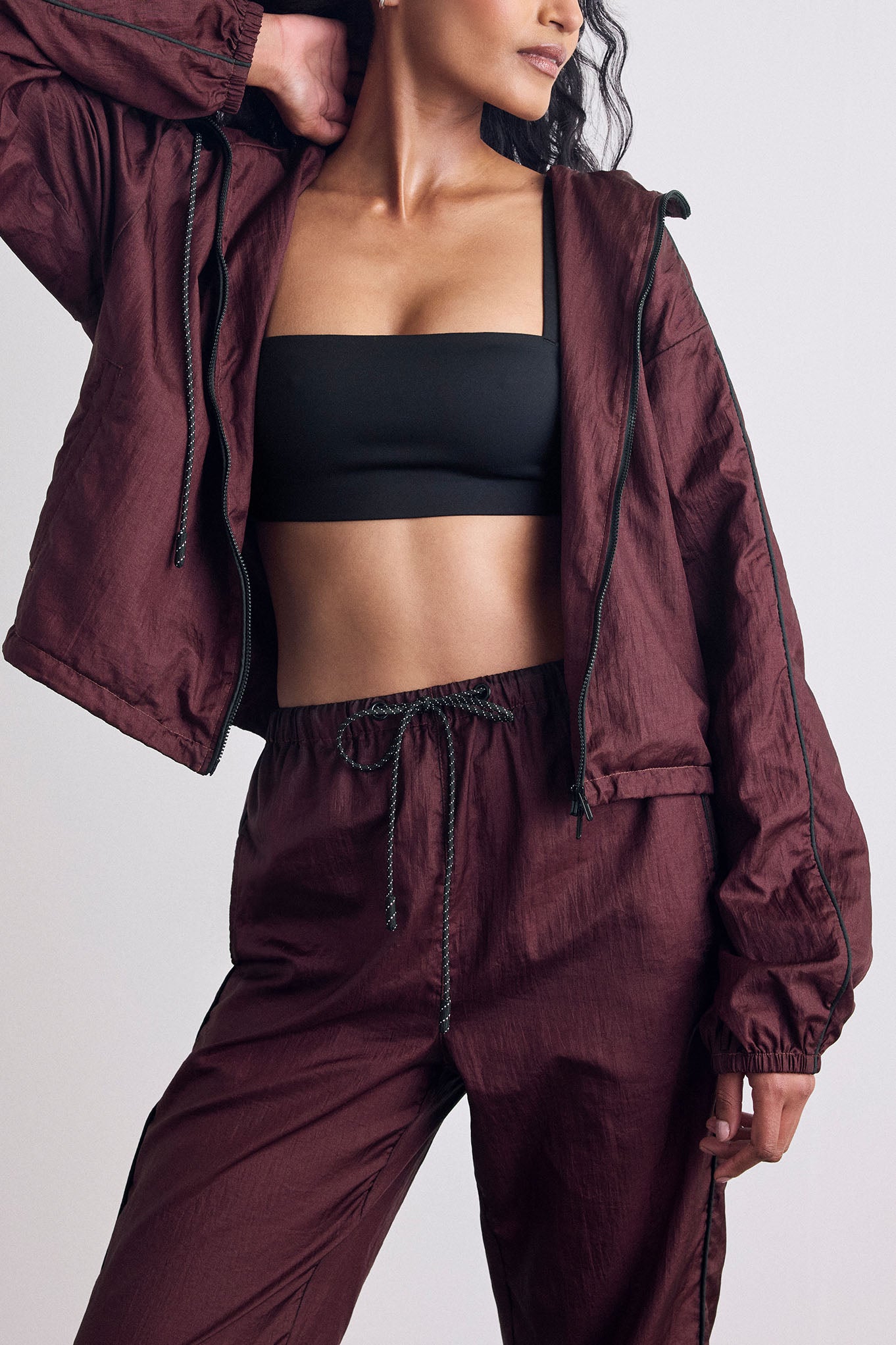 A model wearing a matching burgundy lightweight windbreaker set, featuring an open zip-up jacket and matching drawstring pants, styled over a black sports bra against a light gray studio background.